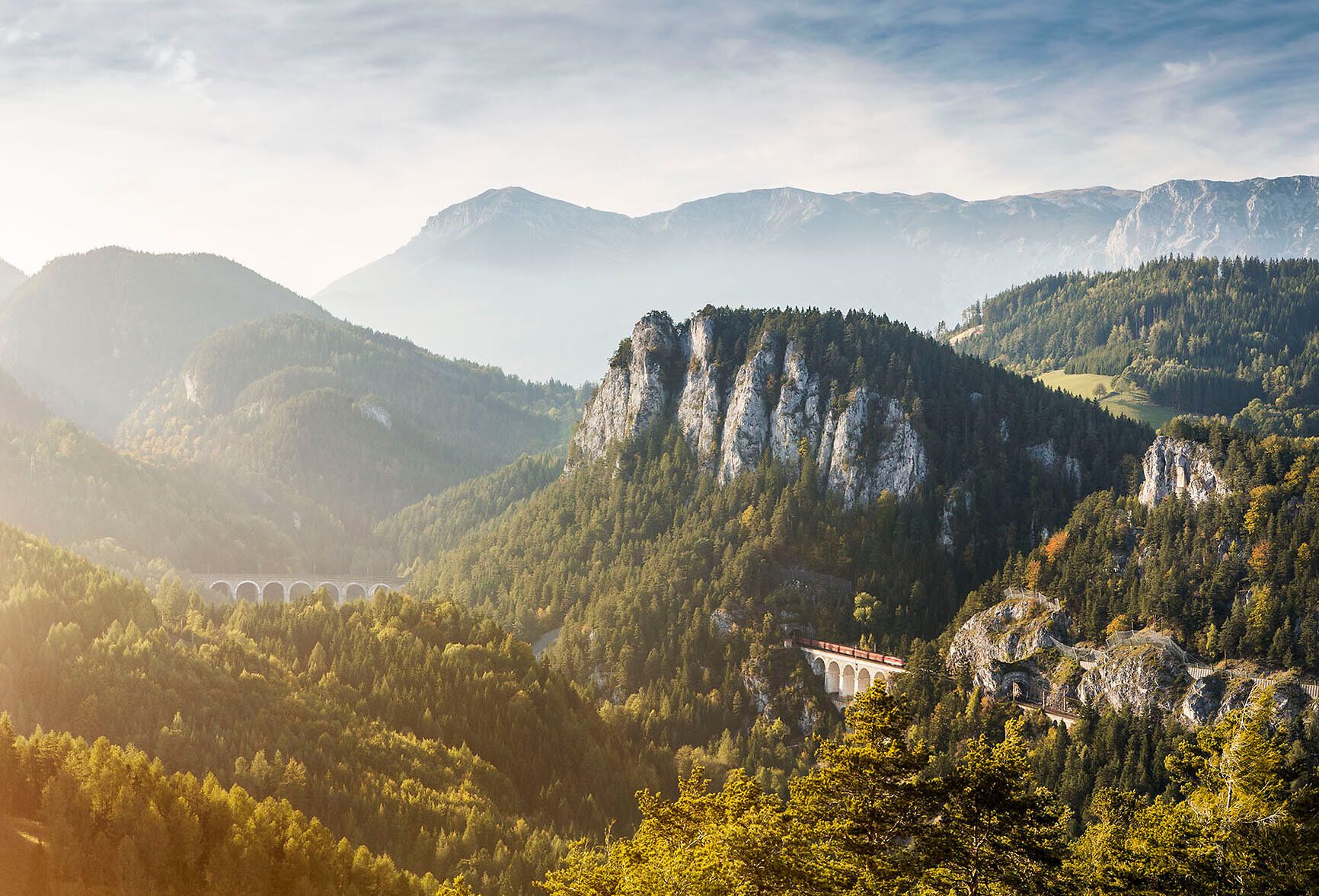 Die sanften Hügel und majestätischen Berge laden zu unvergesslichen Erlebnissen in der Natur ein. Ein sanfter Nebel umhüllt die Landschaft und verleiht der Szenerie eine mystische Atmosphäre, während die Sonne langsam aufgeht und die Farben der Natur zum Leben erweckt.