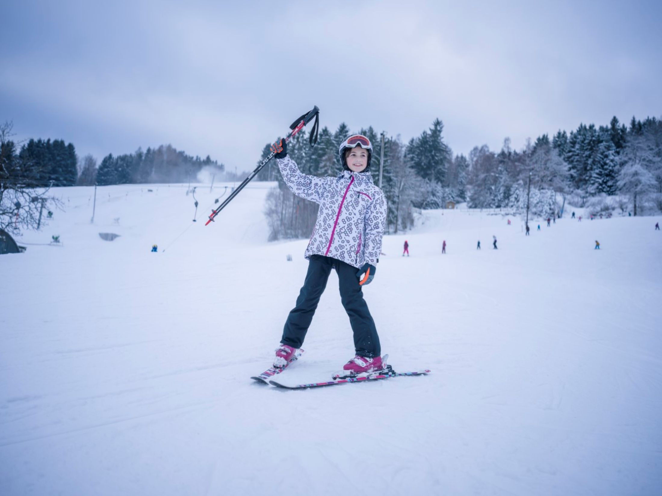 Ein Kind in Skiausrüstung steht auf einer verschneiten Piste und hält Skistöcke in die Luft. Im Hintergrund sind Bäume und andere Skifahrer zu sehen.