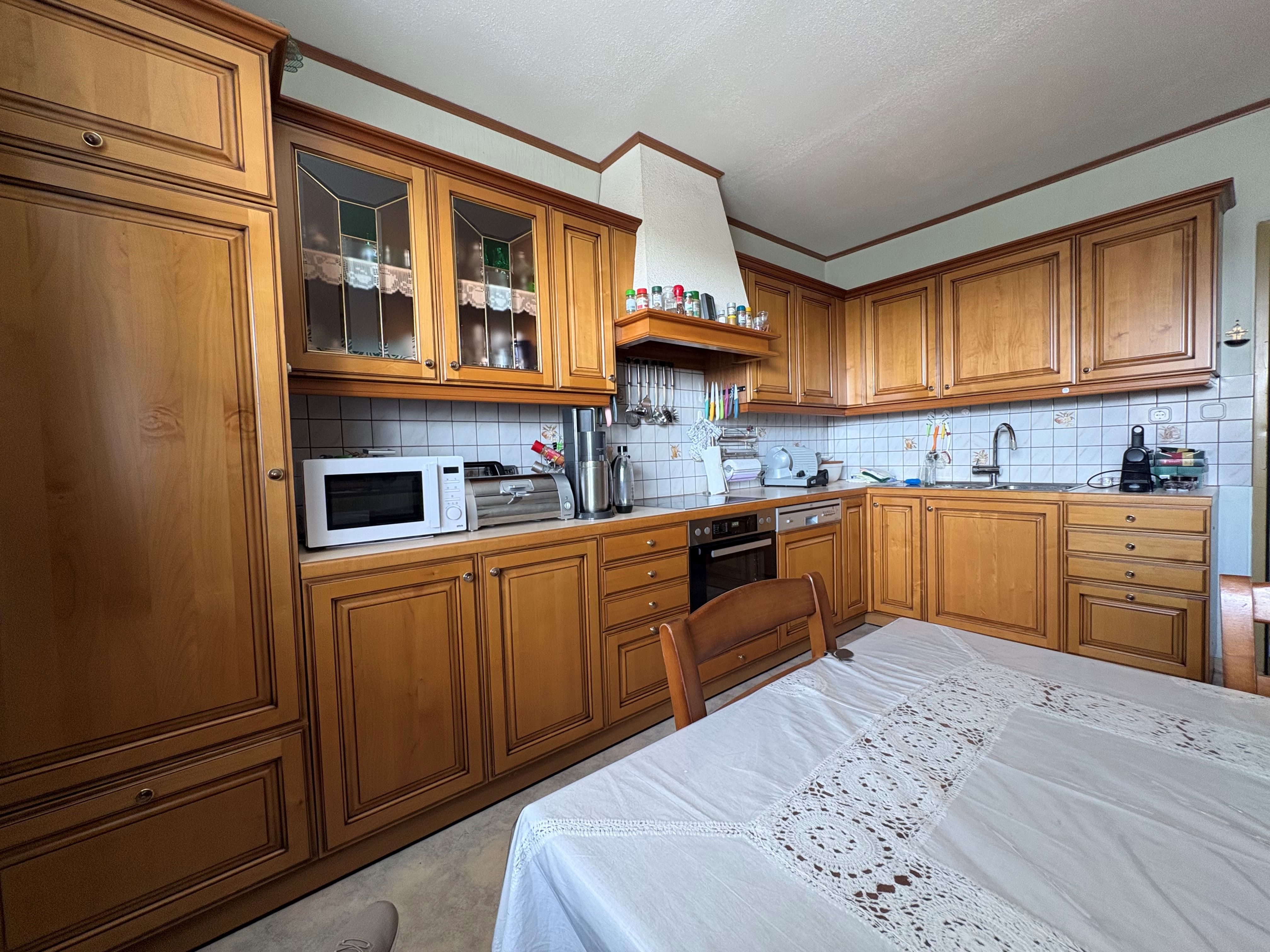 Eine gemütliche Küche mit Holzmöbeln, einem Esstisch mit Spitzentischdecke, Mikrowelle und Küchengeräten auf der Arbeitsplatte.