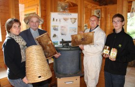 Vier Personen in einem Holzraum mit Imkereiausrüstung und Honiggläsern.