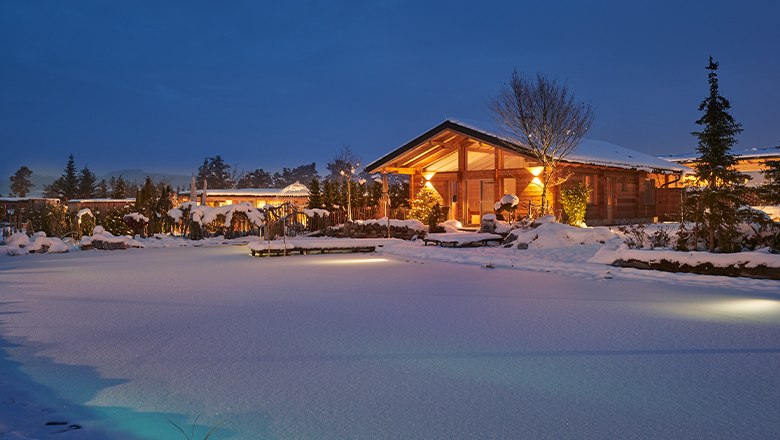 Winterliche Chalets am Gartensee bei Dämmerung, umgeben von Schnee und beleuchtetem Garten.
