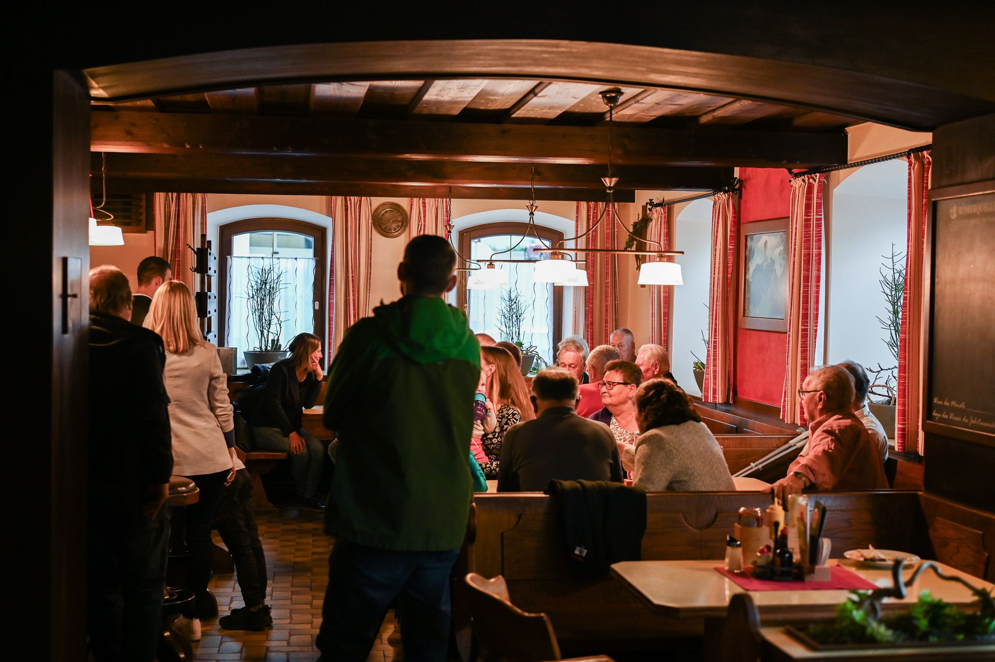 Eine Gruppe von Menschen sitzt in einem gemütlichen, rustikalen Restaurant mit Holzmöbeln und roten Vorhängen.