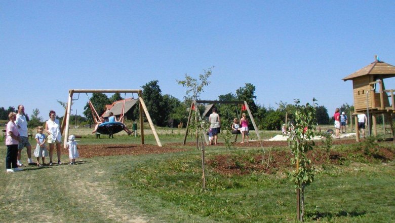 Ein Spielplatz mit Schaukeln, Klettergerüst und Kindern, die spielen. Erwachsene stehen daneben. Es ist ein sonniger Tag.