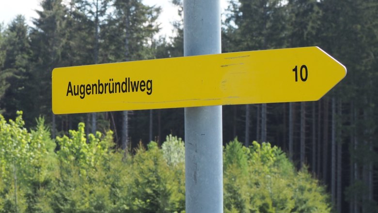 Gelbes Wegweiser-Schild mit der Aufschrift 'Augenbründlweg' und der Zahl '10' vor einem Waldhintergrund.