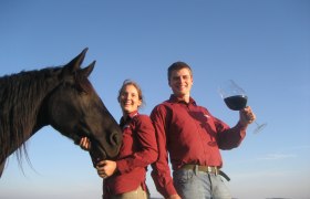 Zwei Personen in roten Hemden mit einem schwarzen Pferd und einem großen Weinglas im Freien.