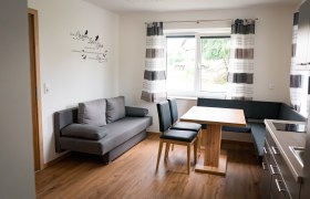 Moderne Küche mit grauem Sofa, Holztisch und Stühlen, Fenster mit Vorhängen.