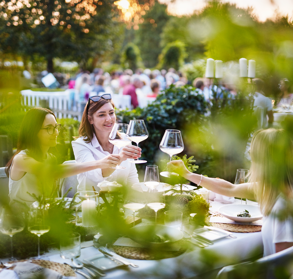 Umgeben von üppigem Grün und der warmen Abendsonne, genießen die Gäste ein geselliges Beisammensein. Die Tische sind liebevoll gedeckt und laden dazu ein, regionale Köstlichkeiten zu probieren und den Sommer in vollen Zügen zu erleben.
