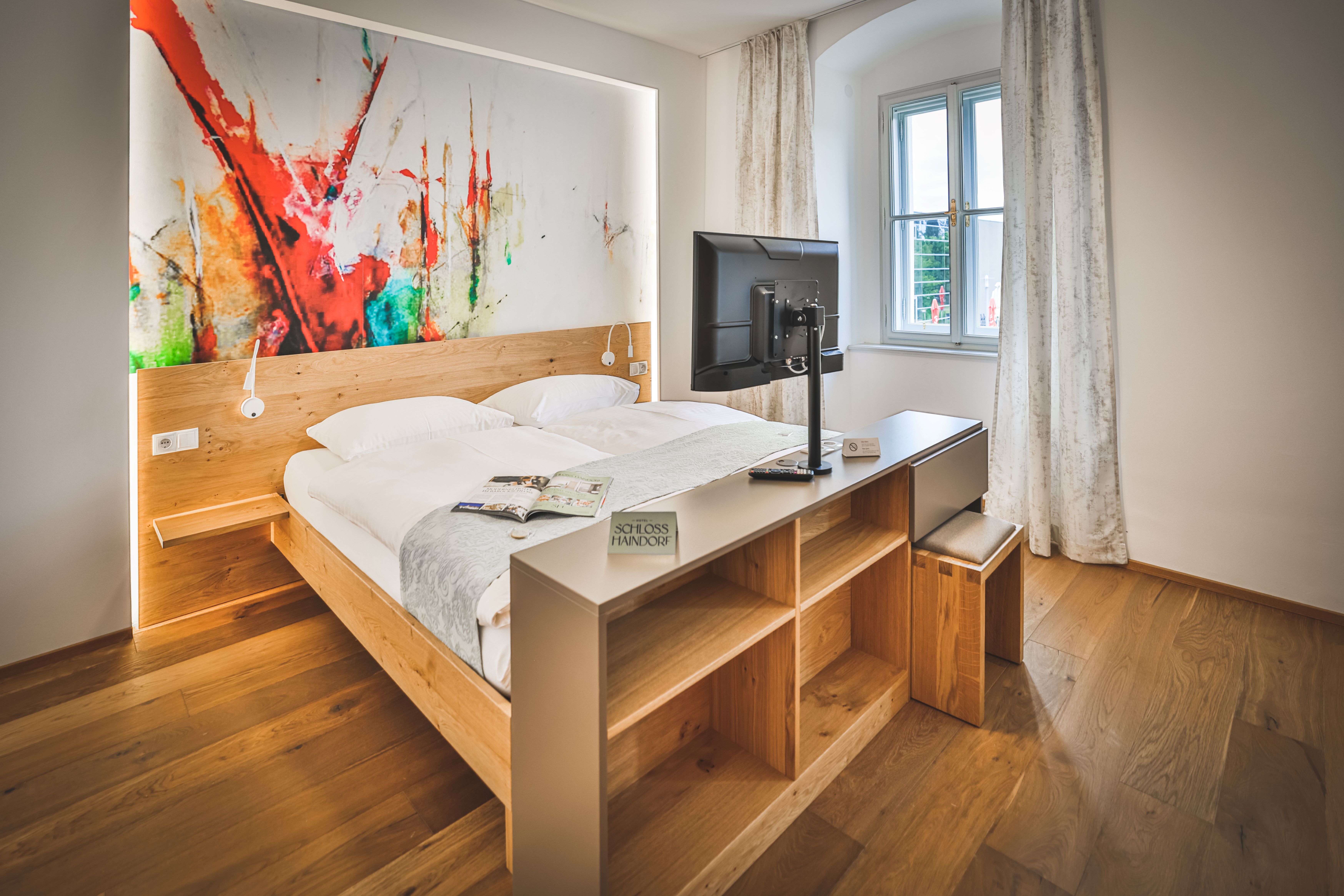 Doppelzimmer im Schloss Haindorf mit Doppelbett, großem abstraktem Wandbild, Fernseher und Fenster mit Vorhängen.