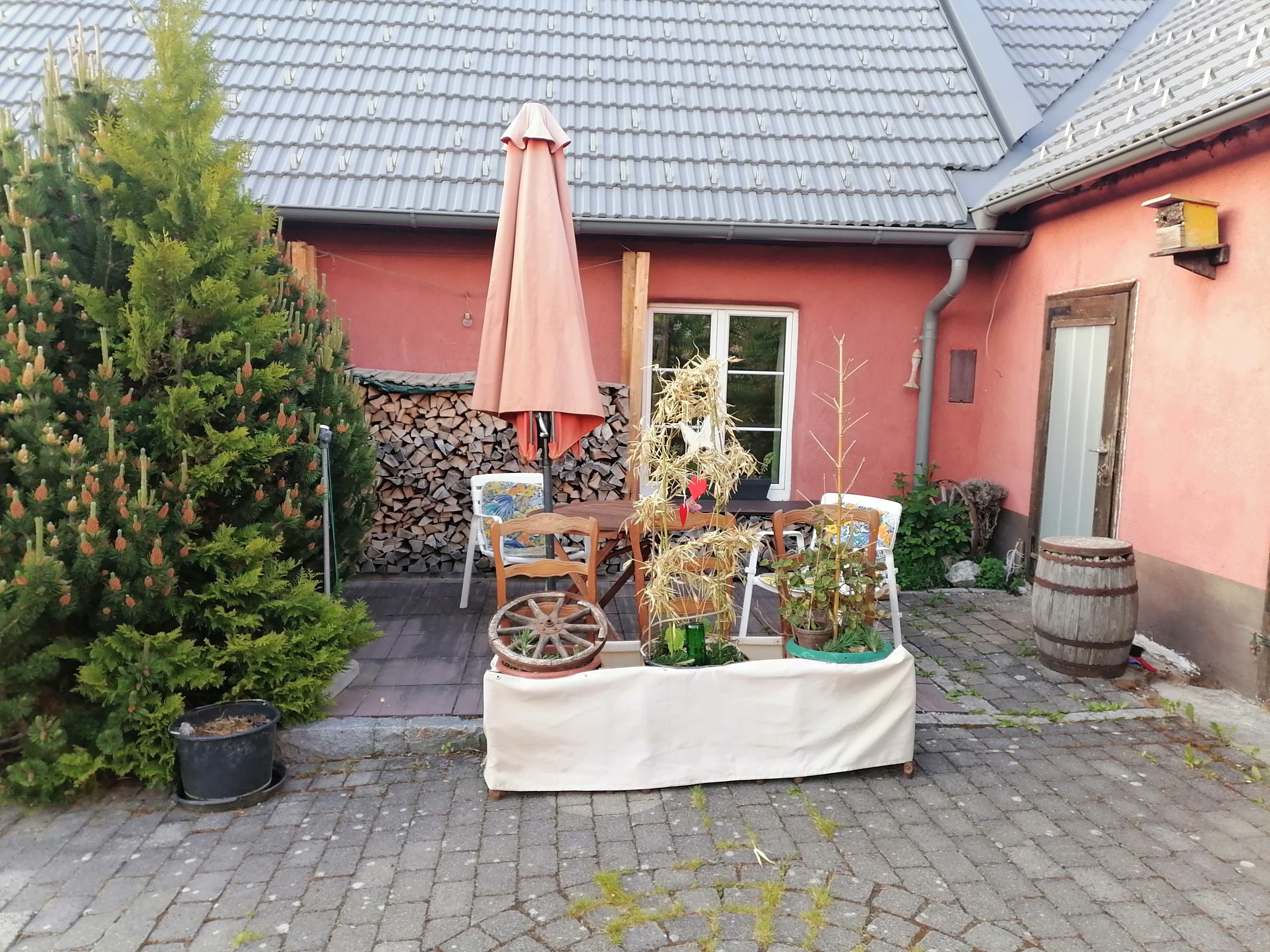 Eine Terrasse mit einem Tisch, Stühlen, einem Sonnenschirm und Pflanzen. Im Hintergrund ein Haus mit Holzstapel und ein großer Baum.