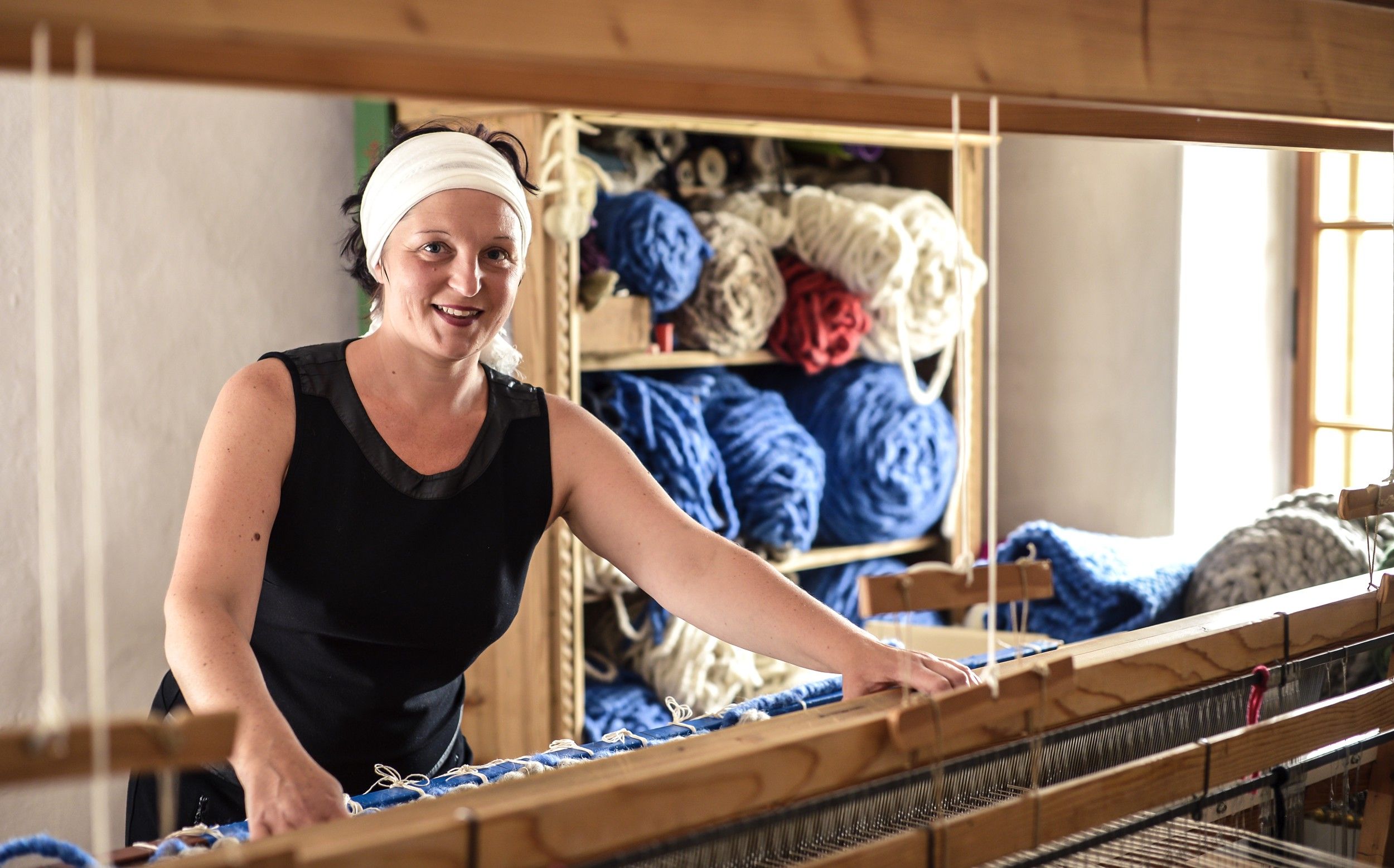 Frau am Webstuhl mit Wollknäueln im Hintergrund.