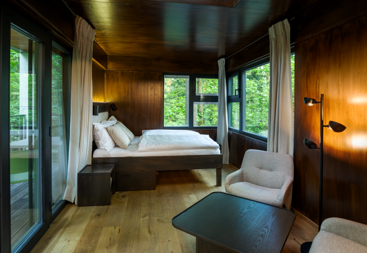 Innenansicht eines modernen Baumhauses mit Holzwänden, Bett, Sessel und großen Fenstern mit Blick ins Grüne.