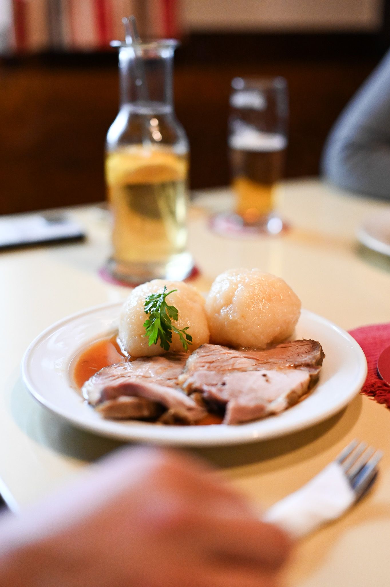 Ein Teller mit Schweinebraten und zwei Knödeln, garniert mit Petersilie. Im Hintergrund ein Glas Bier und eine Karaffe mit Getränk.