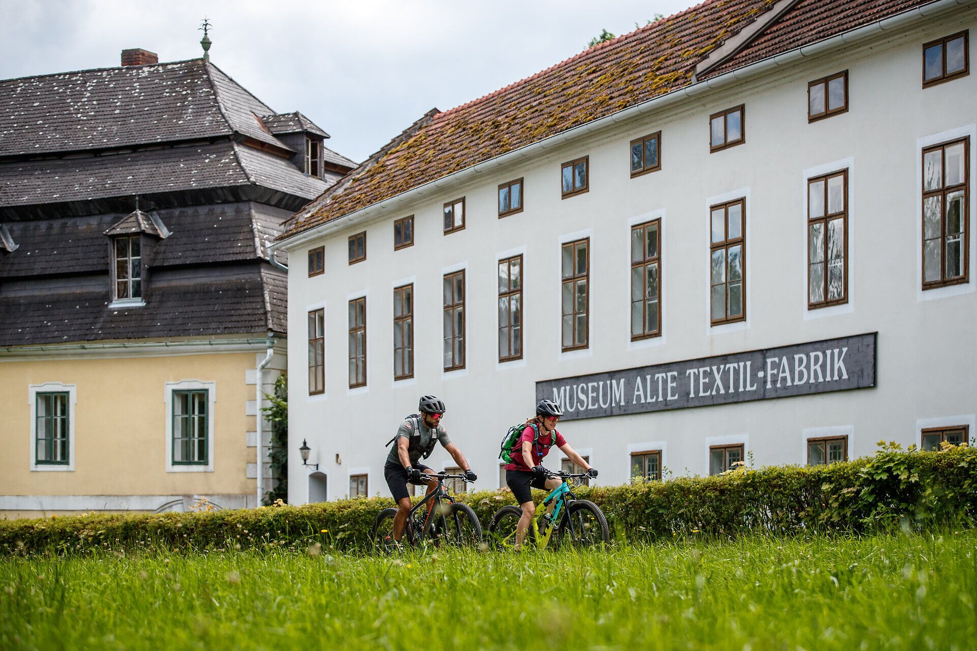 Zwei Mountainbiker gleiten durch die grüne Wiese, umgeben von der beeindruckenden Kulisse historischer Gebäude. Die frische Luft und die sanften Hügel laden dazu ein, die Schönheit der Natur zu genießen und die Freiheit des Radfahrens zu erleben.