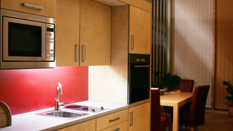 Moderne Küche mit Holzschränken, roter Rückwand, Mikrowelle und Esstisch im Hintergrund.