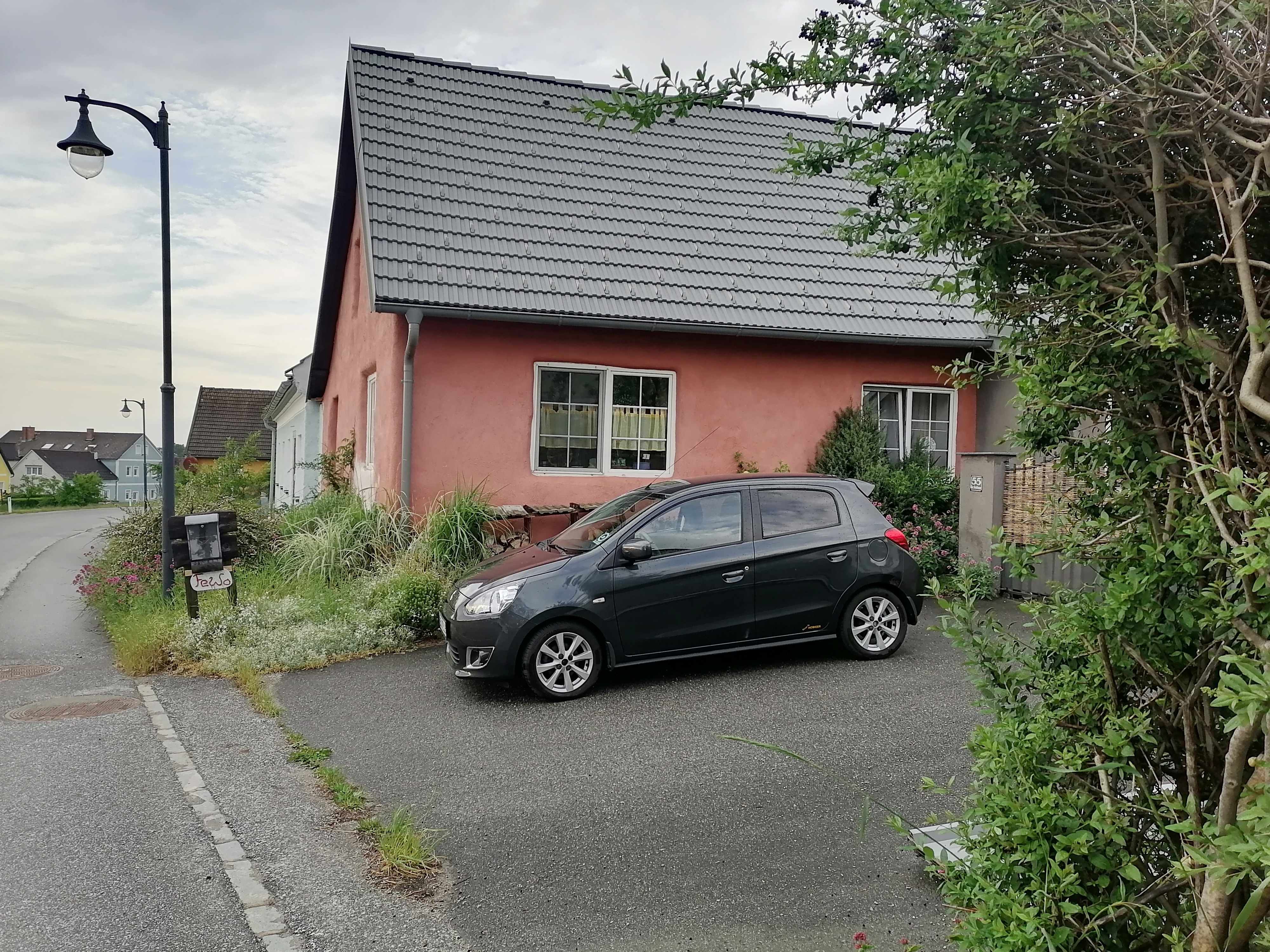 Ein schwarzes Auto parkt vor einem rosa Haus mit grauem Dach. Eine Straßenlaterne steht am Straßenrand.