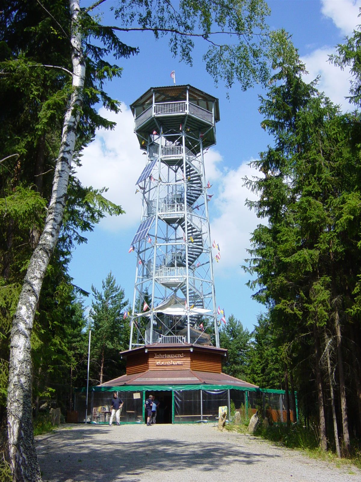 Ein hoher Aussichtsturm aus Metall mit Holzverkleidung, umgeben von Bäumen, unter blauem Himmel.