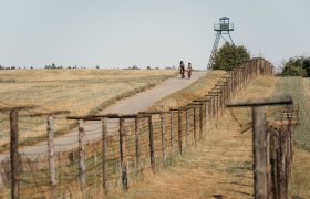 Zwei Radfahrer auf einem Weg neben einem alten Grenzzaun und einem Wachturm.