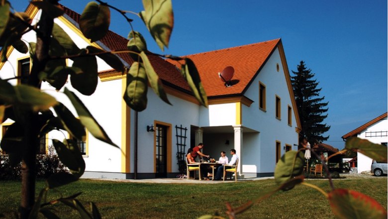 Ferienwohnungen Weixelbaum, © Ferienwohnungen Weixelbaum Ein weißes Haus mit rotem Dach, umgeben von grünem Rasen und Bäumen. Menschen sitzen an einem Tisch im Freien.