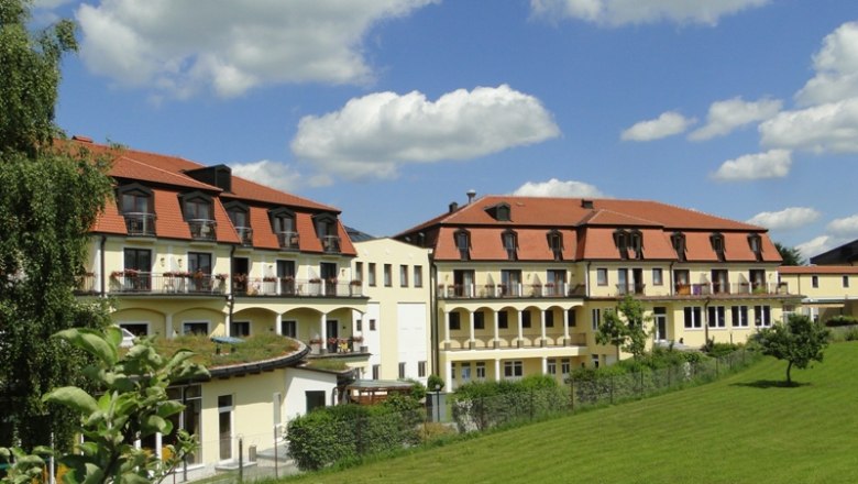 Kurhotel Moorbad Großpertholz mit rotem Dach und Balkonen, umgeben von grüner Wiese und Bäumen unter blauem Himmel.