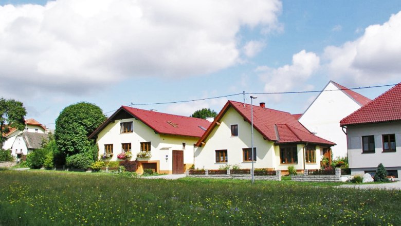 Organic farm Bauer, © Maria Bauer An organic farm with several yellow buildings and red roofs, surrounded by green meadows and trees under a blue sky with clouds.