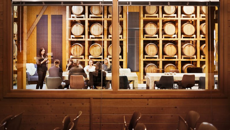 Blick durch ein Fenster in einen Raum mit Whiskyf&auml;ssern und einer Gruppe von Menschen, die an einem Tisch sitzen.