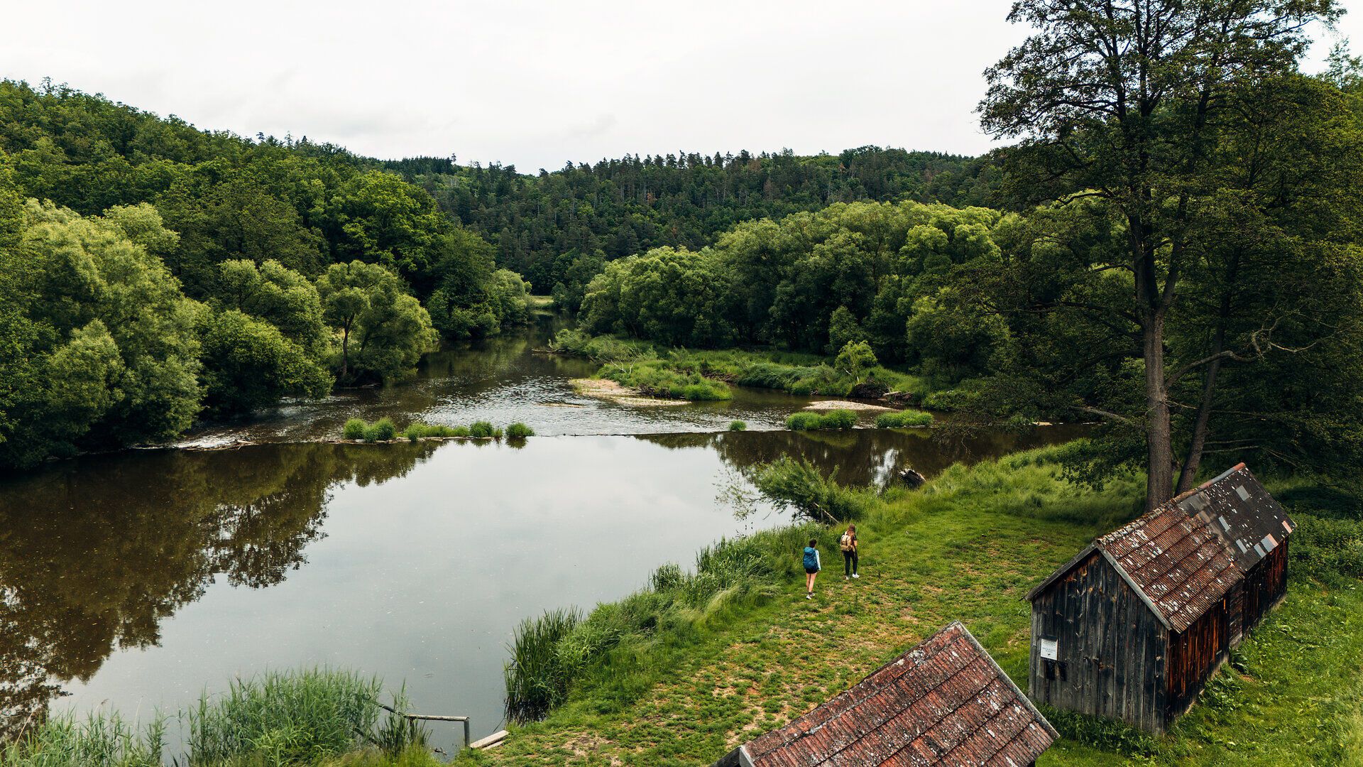 Zwei Personen wandern entlang eines Flussufers, flankiert von dichten Bäumen und alten Holzhütten, in Drosendorf im Waldviertel.
