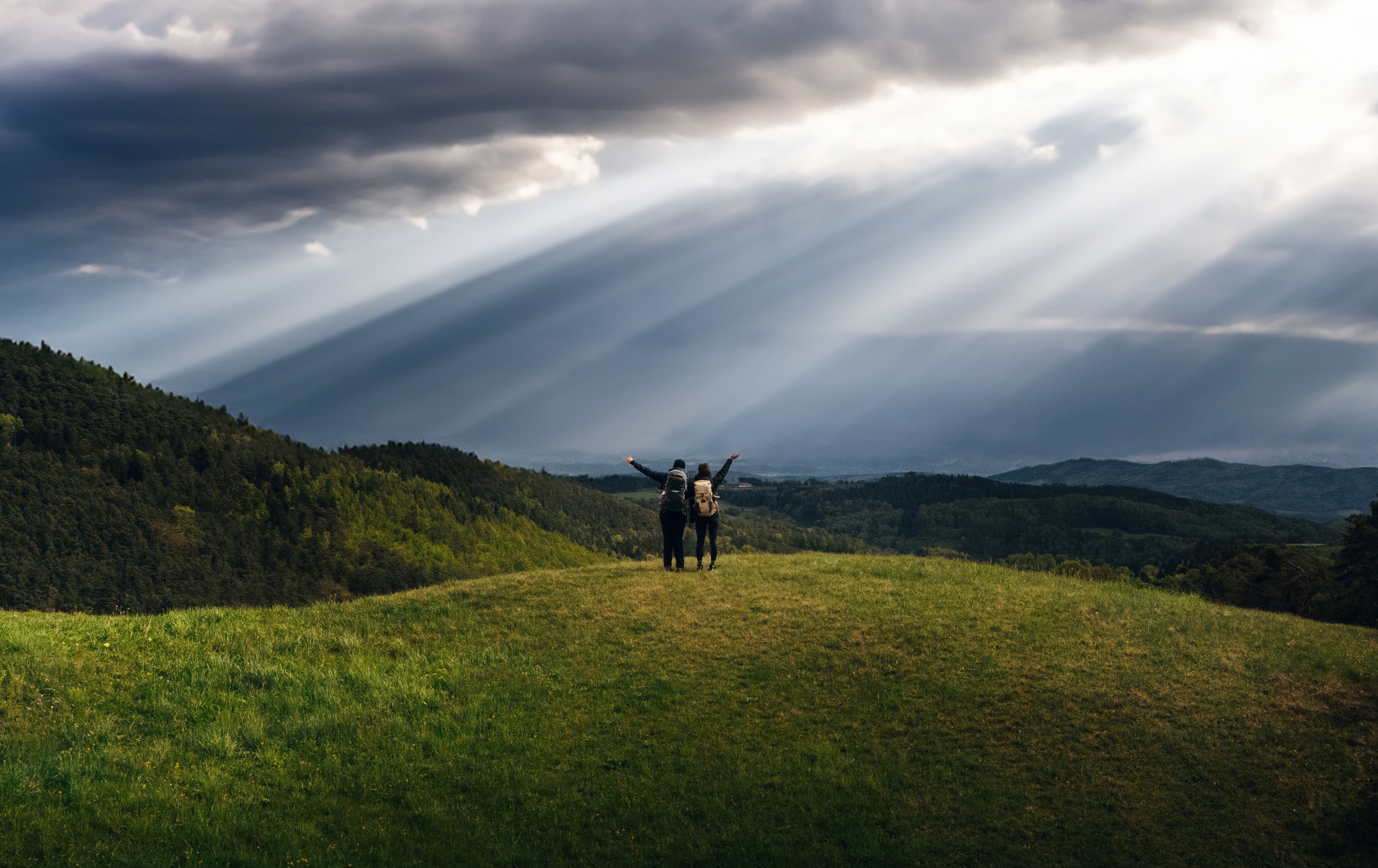 Zwei Personen stehen auf einem Hügel mit Rucksäcken, umgeben von grünen Wäldern und dramatischen Sonnenstrahlen durch Wolken.