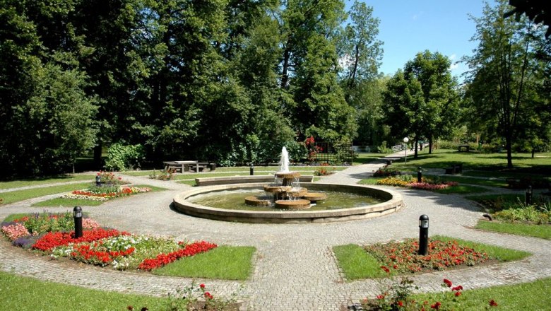 A well-kept park with a central fountain, surrounded by flowerbeds and trees.