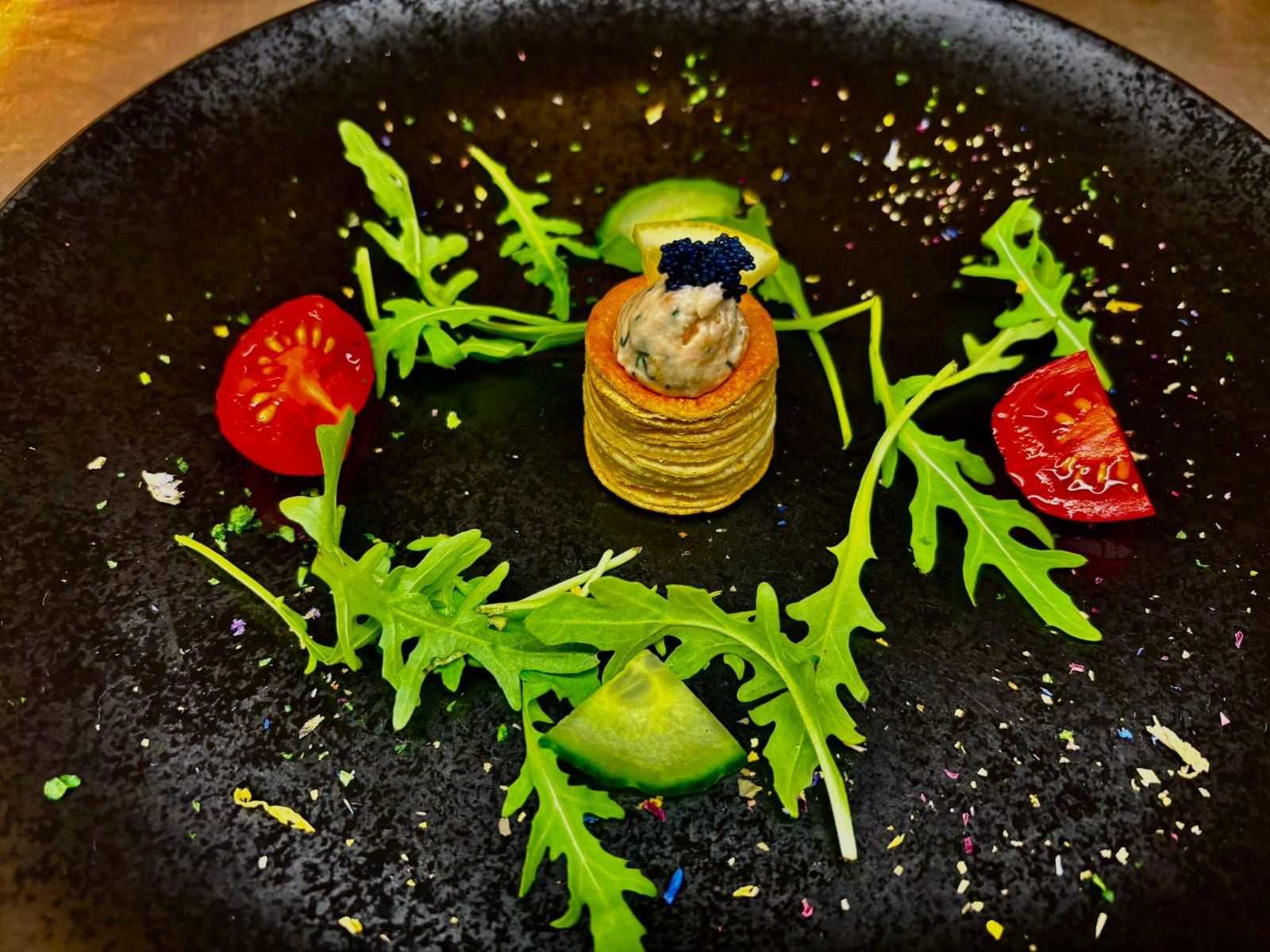 Ein kunstvoll angerichteter Teller mit einem kleinen Stapel Blinis, garniert mit Kaviar, umgeben von Rucola, Tomaten und Gurken auf einem schwarzen Teller.