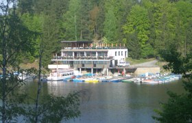 Ein Seerestaurant mit Terrasse und Bootsanleger vor einem Wald.