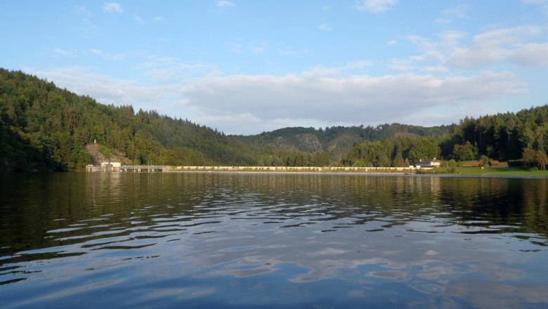 Stausee Thurnberg mit bewaldeten Hügeln und Damm im Hintergrund.