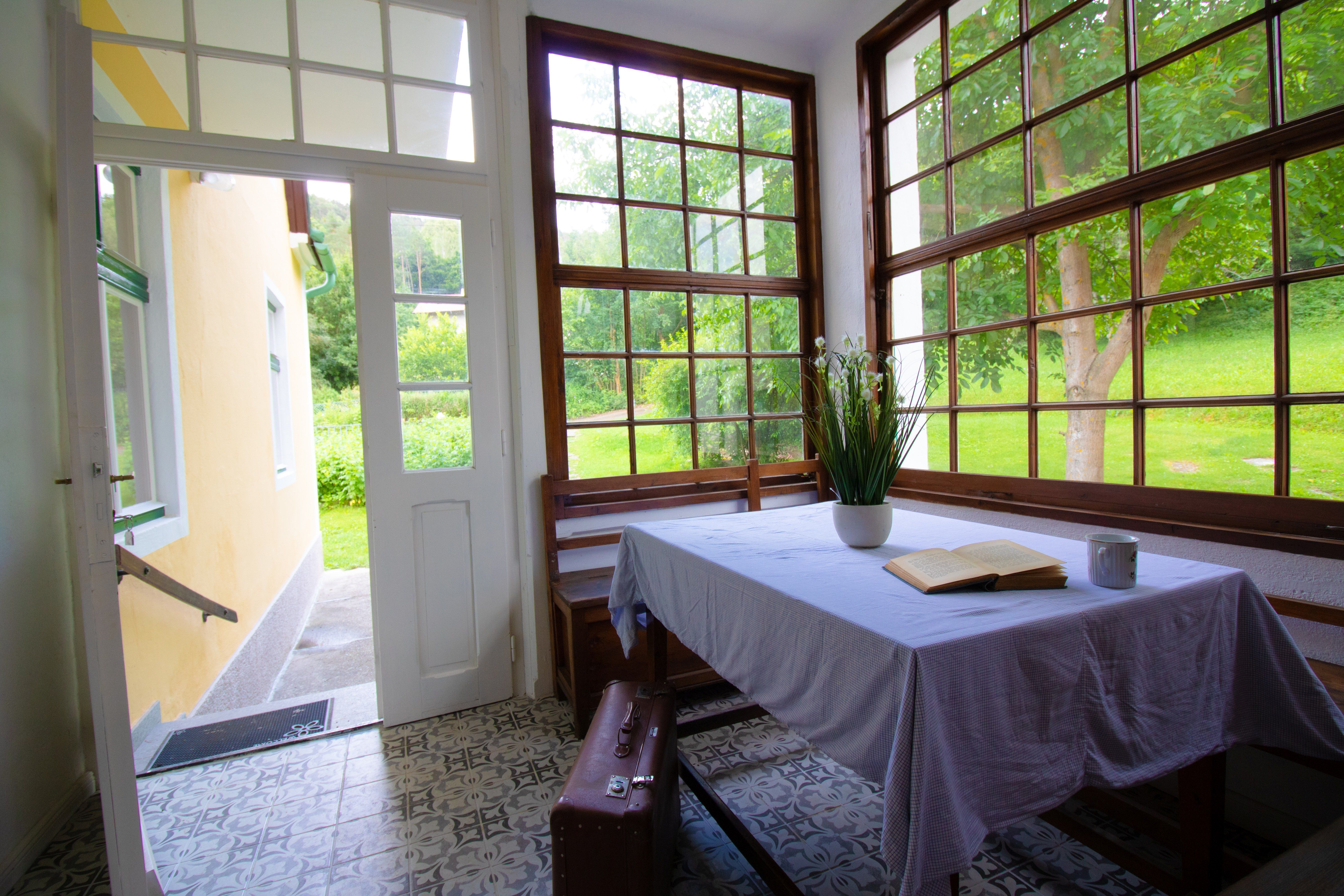 Innenansicht eines hellen Raumes mit Tisch, Buch, Tasse und Pflanzen, umgeben von großen Fenstern mit Blick auf einen Garten.