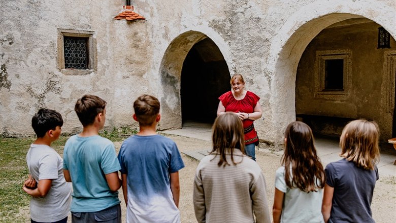 Eine Frau in rotem Oberteil spricht zu einer Gruppe von Kindern vor einer alten Steinmauer mit Torbögen.