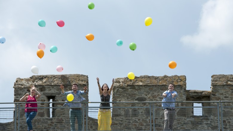 Vier Personen werfen bunte Luftballons von einer Burgmauer in die Luft.