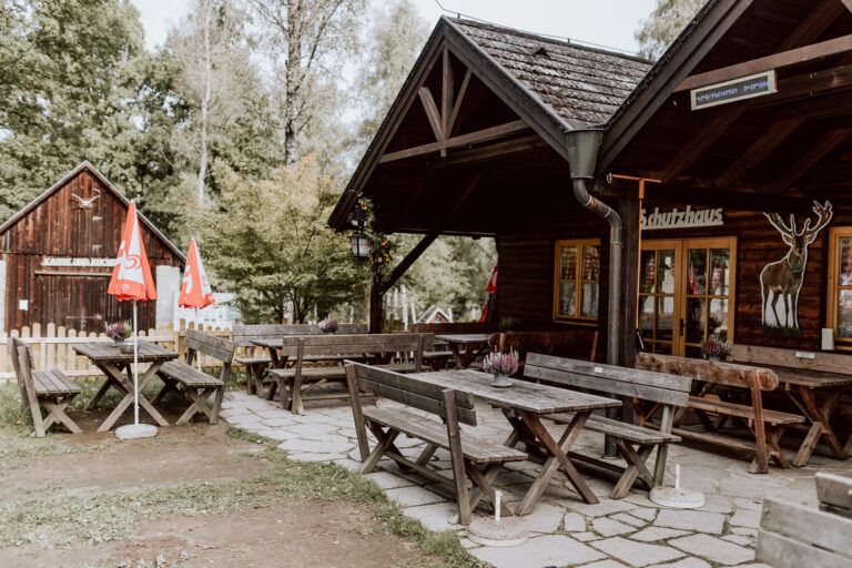 Ein rustikales Gasthaus mit Holzbänken und Tischen im Freien, umgeben von Bäumen und roten Sonnenschirmen.