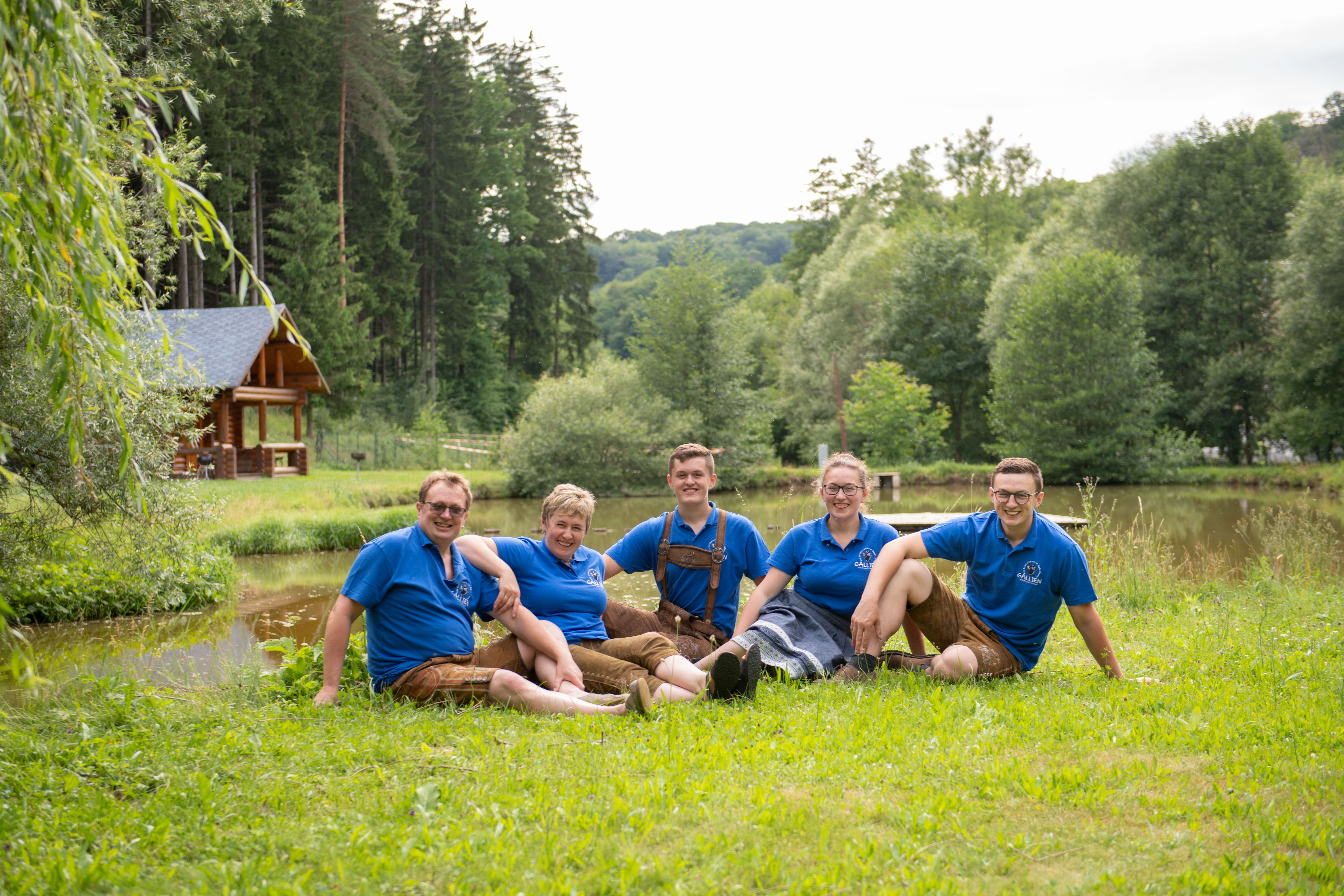 Fünf Personen in blauen Hemden sitzen auf einer Wiese vor einem Teich und einem Holzhaus.
