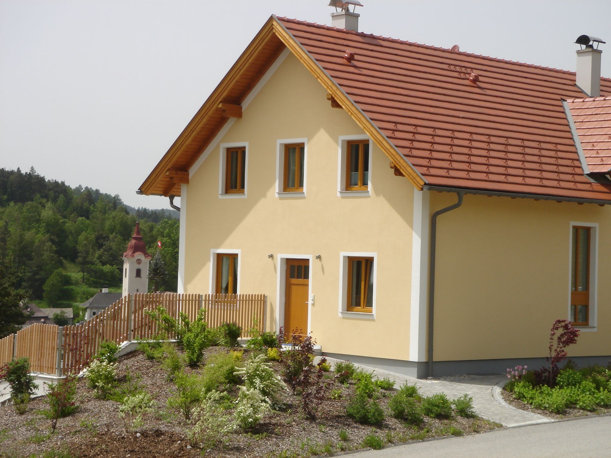 Ein modernes gelbes Haus mit rotem Dach und Garten, im Hintergrund eine Kirche und grüne Landschaft.