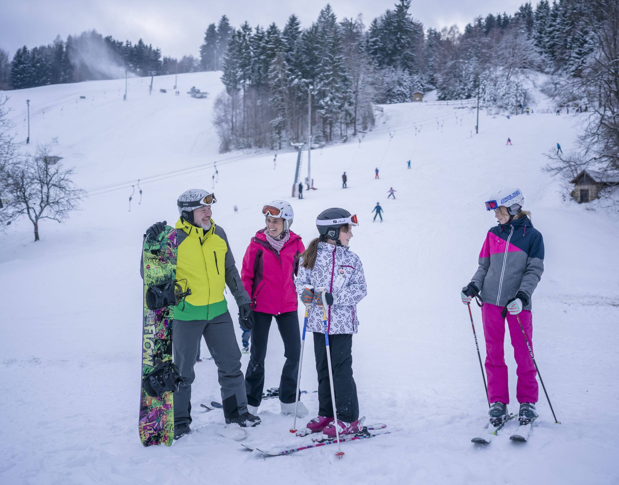 Gruppe von Skifahrern und Snowboardern im Schnee vor einer Skipiste mit Lift.