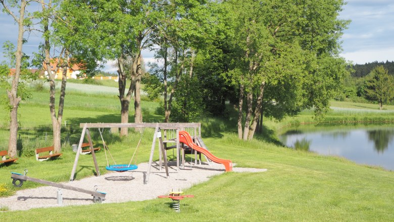 Spielplatz mit Schaukel, Rutsche und Wippe neben einem Teich in einer grünen Landschaft.
