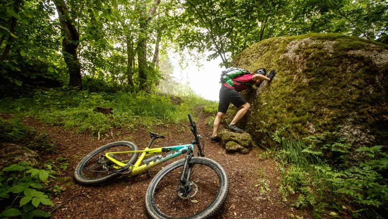 Ein Mountainbiker lehnt an einem großen, moosbedeckten Felsen im Wald, während sein Fahrrad auf dem Boden liegt.