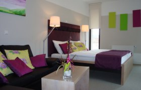 Modern eingerichtetes Hotelzimmer mit Doppelbett, Sofa und Dekoration in Lila- und Grüntönen.