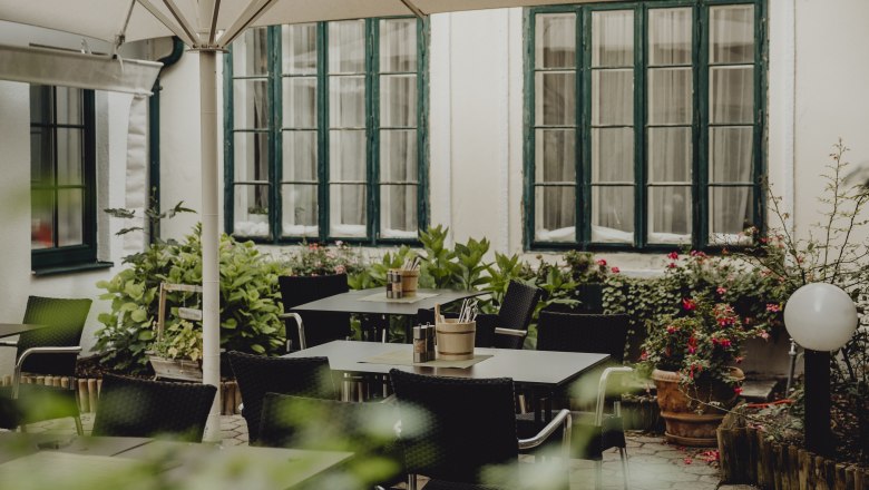 Eine gemütliche Terrasse mit Tischen, Stühlen und Pflanzen, umgeben von großen Fenstern und einem Sonnenschirm.