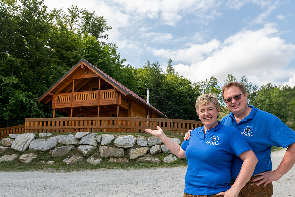 Ein Paar in blauen Polohemden steht vor einer Holzhütte in einer Waldlandschaft.