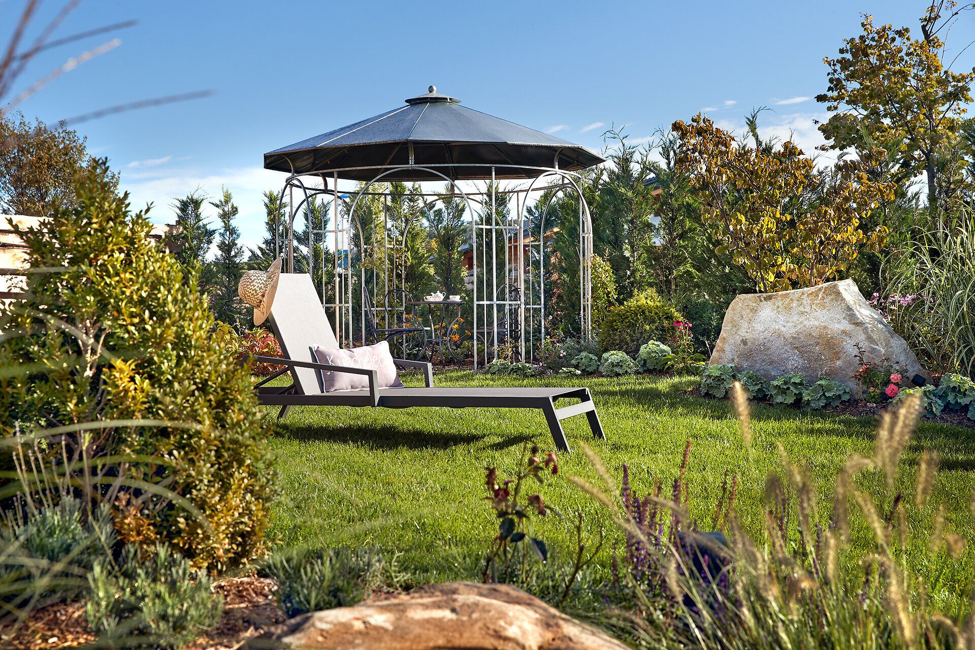 Inmitten einer blühenden Oase lädt eine elegante Liege zum Entspannen ein, während der Blick auf den malerischen Pavillon und die üppige Gartenlandschaft fällt. Die sanften Farben der Pflanzen und das sanfte Licht schaffen eine harmonische Atmosphäre, die zum Verweilen einlädt.