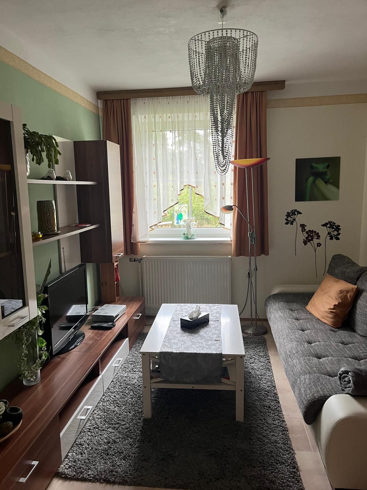 Gemütliches Wohnzimmer mit Sofa, Couchtisch, Teppich und Fernseher. Ein Kronleuchter hängt von der Decke, Pflanzen und Dekorationen sind sichtbar.