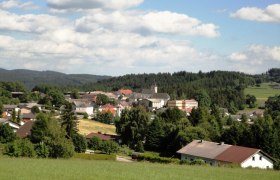 Panorama der Marktgemeinde Martinsberg mit Kirche und umliegenden Häusern, umgeben von Wäldern und Feldern unter blauem Himmel.