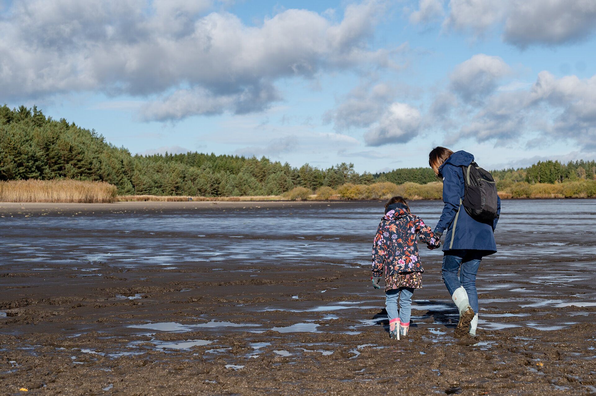 Ein strahlend blauer Himmel spiegelt sich im ruhigen Wasser des Bruneiteichs, während ein Erwachsener und ein Kind Hand in Hand über den schimmernden, schlammigen Boden spazieren. Die sanfte Brise und die umgebende Natur schaffen eine friedliche Atmosphäre, die zum Verweilen einlädt.