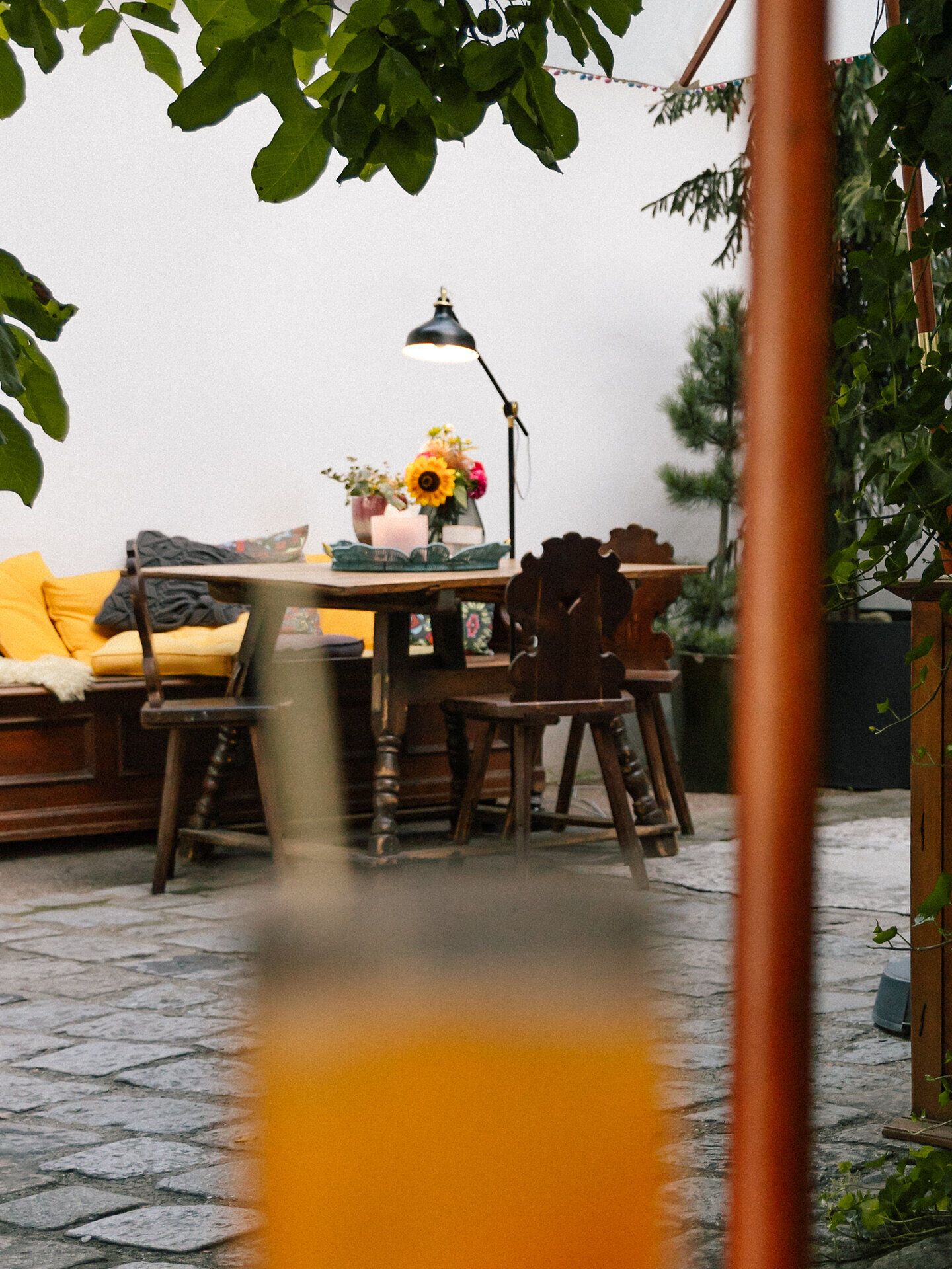 In einem einladenden Gartenbereich laden rustikale Holzmöbel und ein liebevoll gedeckter Tisch mit bunten Blumen zu entspannten Stunden im Freien ein. Die sanfte Beleuchtung des Tischlampen sorgt für eine gemütliche Atmosphäre, während die umgebenden Pflanzen ein Gefühl von Naturverbundenheit vermitteln.