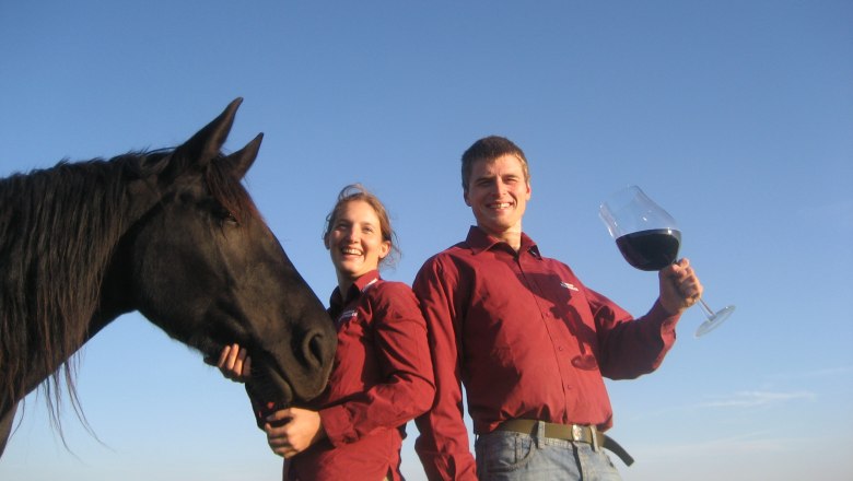 Zwei Personen in roten Hemden mit einem schwarzen Pferd und einem großen Weinglas im Freien.