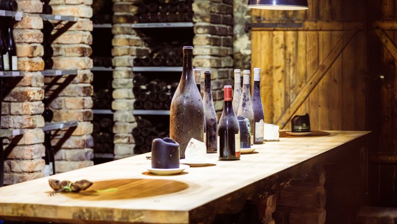 Ein Weinkeller mit Ziegelwänden, Regalen voller Weinflaschen und einem Holztisch mit mehreren Weinflaschen darauf.